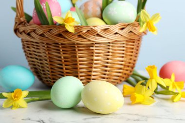 Boyanmış Paskalya yumurtaları ve hasır sepetin içindeki güzel nergisler beyaz mermer masa üzerinde açık mavi arka plan, yakın plan