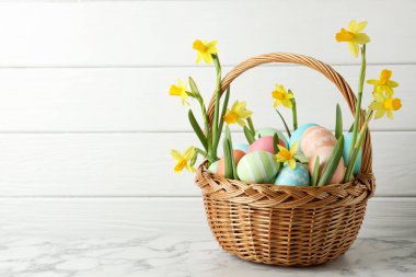 Boyanmış Paskalya yumurtaları ve hasır sepetteki güzel nergisler beyaz mermer masa üzerinde, mesaj için yer var.
