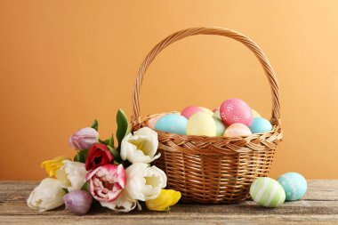 Hasır sepete boyanmış Paskalya yumurtaları ve soluk kahverengi arka plana karşı ahşap masada güzel laleler.