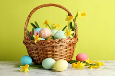 Boyanmış Paskalya yumurtaları ve hasır sepetin içindeki güzel nergisler açık yeşil arka planda beyaz mermer masa üzerinde.