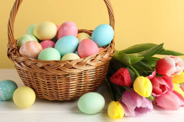 Hasır sepetin içine boyanmış Paskalya yumurtaları ve beyaz ahşap masanın üzerinde güzel laleler. Sarı arka planda, yakın planda.