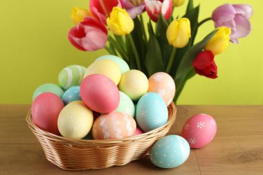 Hasır sepetin içine boyanmış Paskalya yumurtaları ahşap masanın üzerinde ve sarı arka planda güzel laleler, yakın plan.