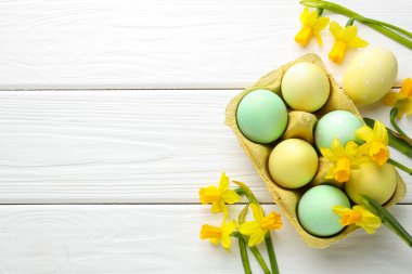 Painted Easter eggs in carton and beautiful daffodils on white wooden table, flat lay. Space for text