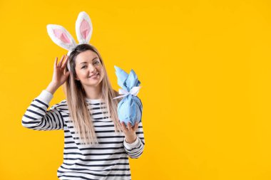 Paskalya kutlaması. Saçbantlı, tavşan kulaklı, sarı arka planda hediyeli gülümseyen kadın, mesaj için yer var.