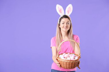 Paskalya kutlaması. Saç bandı takmış, tavşan kulaklı, menekşe arka planda boyalı yumurta dolu sepetli gülümseyen bir kadın.
