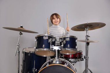 Açık gri arka planda bagetleri ve davulları olan küçük bir çocuk.