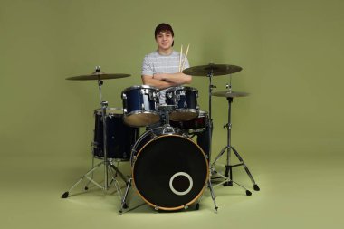 Beyzbol şapkalı, davul setiyle yeşil arka planda gülümseyen genç çocuk.