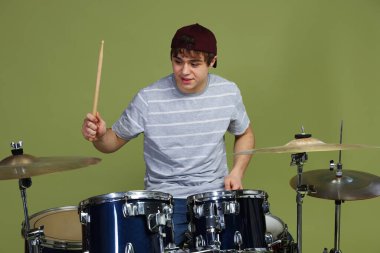 Beyzbol şapkalı, bagetli genç çocuk yeşil arka planda davul çalmayı öğreniyor.