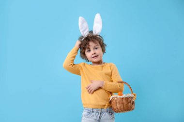 Paskalya kutlaması. Saç bantlı, tavşan kulaklı ve açık mavi arka planda boyalı yumurtalarla dolu sepeti olan sevimli bir çocuk.