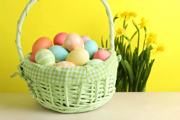Hasır sepetin içine boyanmış Paskalya yumurtaları bej masa üzerinde ve nergisler sarı arka planda, yakın plan