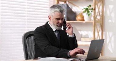 Takım elbiseli duygusal iş adamı akıllı telefonla konuşuyor ve ofiste dizüstü bilgisayarla çalışıyor.