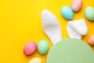 Mutlu Paskalyalar. Şirin tavşan kulakları, boş podyum ve sarı arka planda boyanmış yumurtalar, düz yatak. Metin için boşluk