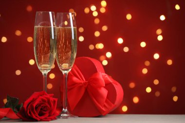 Sevgililer Günü. Köpüklü şarap kadehleri, hediye kutusu ve ahşap masanın üzerinde kırmızı arka planda bulanık ışıklar ve yazı için yer olan gül. Bokeh etkisi
