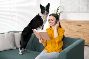 Kulaklıklı gülümseyen kadın kitap okuyor ve evdeki kanepede sevimli köpeği oturuyor.