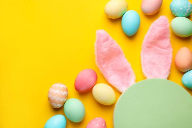 Mutlu Paskalyalar. Şirin tavşan kulakları, boş podyum ve sarı arka planda boyanmış yumurtalar, düz yatak. Metin için boşluk