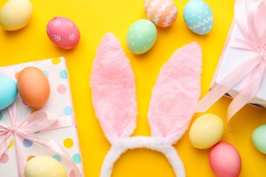 Boyalı Paskalya yumurtaları, tavşan kulaklıkları ve sarı arka planda hediyeler.