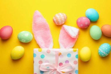 Boyalı Paskalya yumurtaları, tavşan kulaklıkları ve sarı arka planda hediyeler, düz yatış.