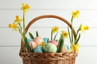Boyanmış Paskalya yumurtaları ve hasır sepetin içindeki güzel nergisler beyaz ahşap arka planda, yakın plan.