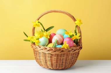 Boyanmış Paskalya yumurtaları ve hasır sepetin içindeki güzel nergisler bej arka planda beyaz ahşap masa üzerinde