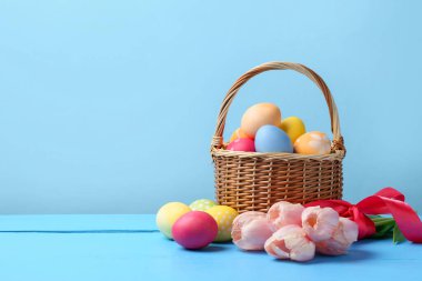 Hasır sepete boyanmış Paskalya yumurtaları ve açık mavi arka plana karşı ahşap masada güzel laleler. Metin için boşluk