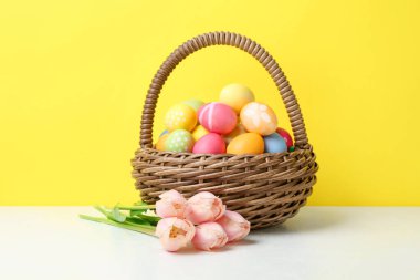 Hasır sepette boyanmış Paskalya yumurtaları ve sarı arka planda beyaz masada güzel laleler.