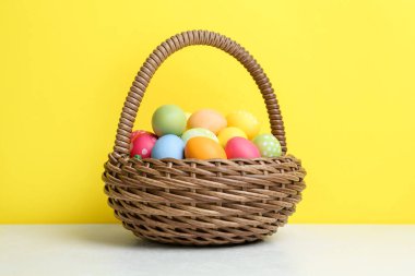 Hasır sepetin içine boyanmış Paskalya yumurtaları beyaz masada sarı arka planda.