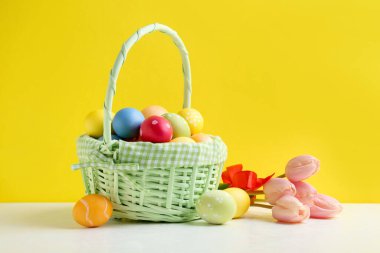 Hasır sepette boyanmış Paskalya yumurtaları ve sarı arka planda beyaz masada güzel laleler.