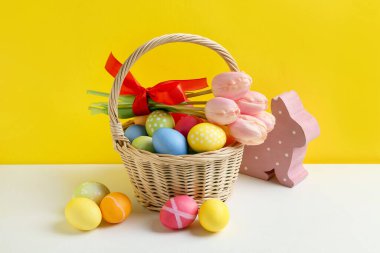 Hasır sepette güzel lalelerle boyanmış Paskalya yumurtaları ve sarı arka planda beyaz masa üzerinde dekoratif tavşancık.