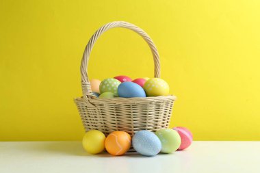 Hasır sepetin içine boyanmış Paskalya yumurtaları beyaz masada sarı arka planda.