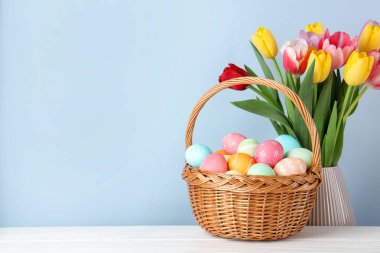 Hasır sepetin içine boyanmış Paskalya yumurtaları beyaz ahşap masa üzerinde ve açık mavi arka planda vazoda güzel laleler. Metin için boşluk