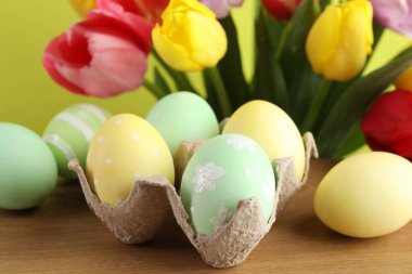 Ahşap masada karton içine boyanmış Paskalya yumurtaları ve sarı arka planda güzel laleler. Yakın plan.
