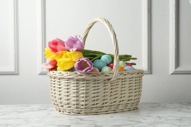 Hasır sepete boyanmış Paskalya yumurtaları ve beyaz duvarın yanındaki beyaz mermer masada güzel laleler.