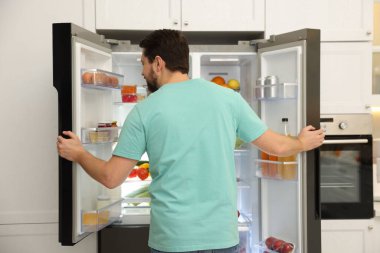 Mutfakta farklı gıda ürünleri olan açık buzdolabının yanındaki adam.