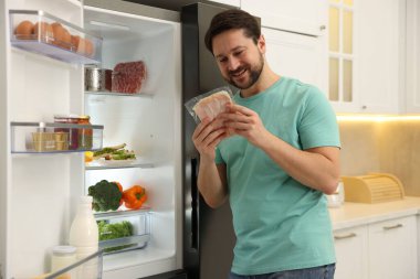 Buzdolabının yanında jambon dilimleri olan adam mutfaktaki yiyeceklerle dolu.