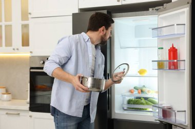 Mutfaktaki açık buzdolabının yanında tavası ve kapağı olan bir adam.