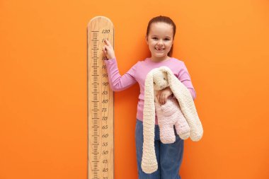 Oyuncak tavşancığı olan küçük kız turuncu duvarın yanında boyunu ölçüyor.