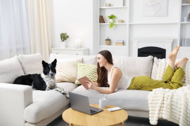 Sevimli köpeğiyle evdeki kanepede kitap okuyan gülümseyen kadın.