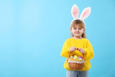 Paskalya kutlaması. Saç bandı takmış, tavşan kulaklı, açık mavi arka planda boyalı yumurta dolu sepet tutan şirin bir kız portresi.