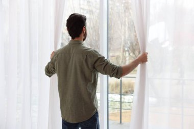 Adam evde, arka planda şık pencere perdeleri açıyor