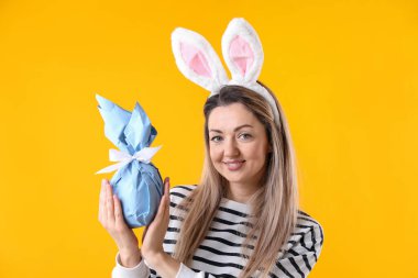 Paskalya kutlaması. Saç bandı takmış, tavşan kulaklı, gülümseyen bir kadın sarı arka planda hediyesini tutuyor.