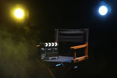Film yönetmeni koltuğu, alkış tahtası ve siyah arka plandaki ışıklandırma ekipmanları dumanlı.