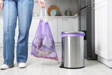 Mutfakta plastik poşet dolusu çöp taşıyan bir kadın var.