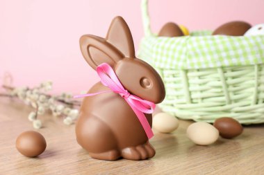 Çikolatalı Paskalya tavşanı ve hasır sepetteki yumurtalar ahşap masada pembe arka planda, yakın plan.
