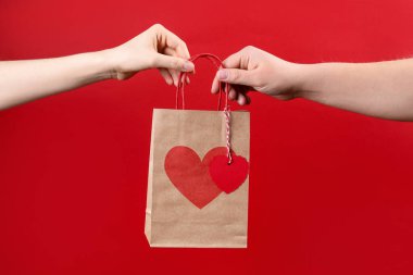Erkek, kırmızı arka planda sevgililer günü hediyesi olan kağıt torbayı kadına veriyor.