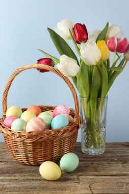 Hasır sepete boyanmış Paskalya yumurtaları ve ahşap masada açık mavi arka planda güzel laleler. Yakın plan.