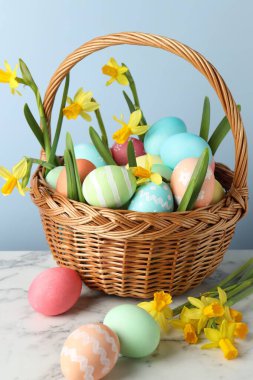 Boyanmış Paskalya yumurtaları ve hasır sepetin içindeki güzel nergisler beyaz mermer masa üzerinde açık mavi arka plan, yakın plan