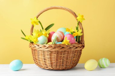 Boyanmış Paskalya yumurtaları ve hasır sepetin içindeki güzel nergisler bej arka planda beyaz ahşap masa üzerinde
