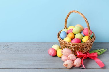 Hasır sepete boyanmış Paskalya yumurtaları ve açık mavi arka planda gri ahşap masada güzel laleler. Metin için boşluk