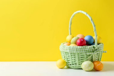 Hasır sepetin içine boyanmış Paskalya yumurtaları beyaz masada, sarı arka planda. Metin için boşluk