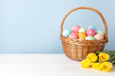Hasır sepete boyanmış Paskalya yumurtaları ve açık mavi arka planda beyaz ahşap masada güzel laleler. Metin için boşluk